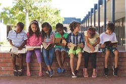 Front view of diverse students reading book while sitting on brick wall at corridor in school