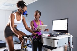 Front view of an African-American athletic woman doing a fitness test using a mask connected to a monitor while riding an exercise bike and an African-American woman monitoring her inside a room at a