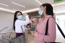 Two mixed race female colleagues working in a modern office wearing face masks greeting each other by touching elbows. Hygiene and social distancing in workplace during Coronavirus Covid 19 pandemic.