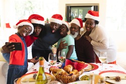 Multigeneration family wearing santa hats making faces while taking a selfie from smartphone while standing near the dining table in the living room at home.
