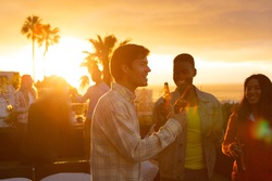 Front view of a multi-ethnic group of friends hanging out on a roof terrace with a sunset sky, holding bottles of beer and smiling