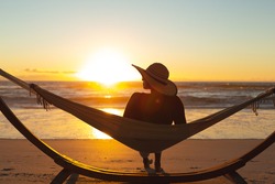 Mixed race woman on beach holiday sitting in hammock during sunset. healthy outdoor leisure time by the sea.