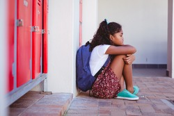 Unhappy african american schoolgirl sitting by lockers in school corridor with schoolbag. childhood and education at elementary school.