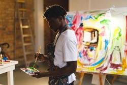 African american male painter at work holding paints and brush in art studio. creation and inspiration at an artists painting studio.