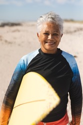 Portrait of smiling senior biracial woman with short white hair holding surfboard at sunny beach. water sport and active lifestyle.