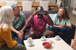Happy multiracial senior male and females looking at friend using vr headset at home. unaltered, lifestyle, social gathering, friendship, virtual reality, futuristic and modern technology.