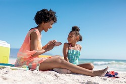 Happy african american woman applying suntan lotion on daughter while sitting at beach on sunny day. unaltered, family, lifestyle, togetherness, enjoyment and holiday concept.