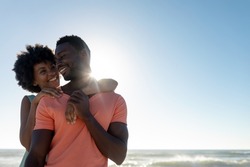 Happy back lit african american woman embracing boyfriend at beach against blue sky with copy space. unaltered, lifestyle, love, togetherness and holiday concept.