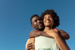 Low angle view of smiling african american man hugging girlfriend from behind against blue sky. unaltered, copy space, lifestyle, love, togetherness and holiday concept.
