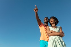 Low angle view of happy african american man pointing with girlfriend against clear blue sky. unaltered, copy space, lifestyle, love, togetherness and holiday concept.