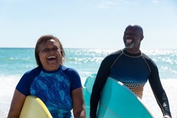 Cheerful african american senior retired couple carrying surfboards enjoying sunny day at beach. unaltered, togetherness, active lifestyle, aquatic sport and holiday concept.