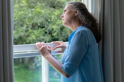 Contemplative caucasian senior woman looking away while standing at window in house. unaltered, lifestyle, retirement, contemplation, loneliness.