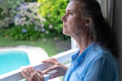 Contemplative caucasian senior woman looking away while standing at window. unaltered, lifestyle, retirement, contemplation, loneliness.