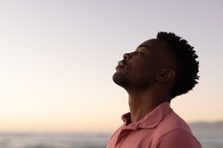 Side view of african american young man with eyes closed at beach against clear sky during sunset. copy space, nature, unaltered, beach, lifestyle, enjoyment and holiday concept.