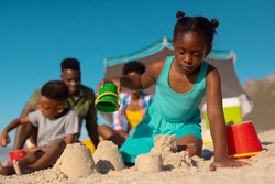 Low angle view of african american girl making sand castle against sky with family in background. copy space, unaltered, beach, childhood, family, togetherness, lifestyle, enjoyment, holiday.