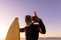 Portrait of bearded african american senior man with surfboard gesturing at beach against clear sky. copy space, happy, unaltered, retirement, aquatic sport, holiday and active lifestyle concept.