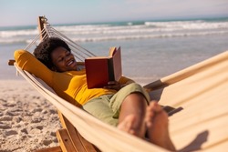 Smiling african american mature woman reading book while lying on hammock against sea in summer. 