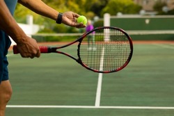 Midsection of biracial senior man holding racket serving tennis ball while playing at tennis court. unaltered, sport, competition, retirement, healthy and active lifestyle concept.