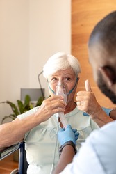 Caucasian senior woman showing thumbs up while using oxygen mask sitting on a wheelchair at home. Medical care and retirement senior lifestyle concept
