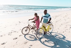 High angle view of african american senior couple with bikes talking and walking at beach in summer. Copy space, travel, unaltered, vacation, love, together, retirement, nature and active lifestyle.