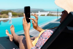 African american wearing straw hat, sitting on deckchair and using smartphone by swimming pool. Spending quality time at home alone, domestic life and lifestyle concept.
