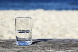 Glass of water which is half-full stands on a wooden table which stands on the sand beach by the sea