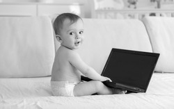 Black and white image of 1 year old baby boy sitting on sofa and using laptop