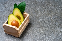 Avocado in wooden box with leaves on gray background.