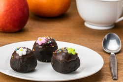 Choc balls or chocolate balls topping with multicolored rainbow sprinkles and colored sugar flower in white ceramic dish on wooden table with red apple, ripe orange and a cup of coffee.