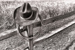 American West rodeo traditional cowboy hat and roping lasso lariat hanging on an old wood fence post near a western ranch field in black and white