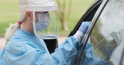As part of the operations of a coronavirus mobile testing unit a healthcare worker dressed in full protective gear swabs an unseen person sitting inside of a vehicle.