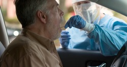 A medical technician in full protective gear collects a sample from a mature man sitting inside his car as part of the operations of a coronavirus mobile testing unit.