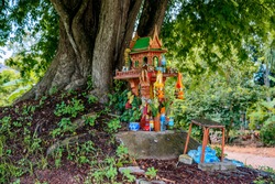 A spirit house on earth mound with huge tree stands behind. Traditional Thai Miniature house built for guardian spirit to reside. Food and drink are common offering from Buddhists and worshipers.