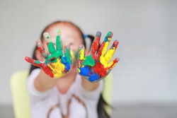 Portrait of smiling little girl looking through her colorful hands and cheek painted in kids room. Focus at baby hands.