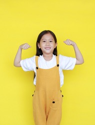 Strong asian little kid girl raising hands up and smiling. Portrait of happy child in dungarees isolated on yellow background.