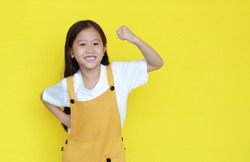 Happy asian little child girl showing his muscle with looking camera isolated on yellow background with copy space. Strong kid in dungarees with good health concept