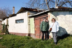 Two civilians standing in front of an old neglected house