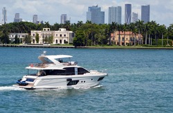 White motor yacht cruising by luxury homes on Star Island in Miami Beach,Florida with downtown Miami tall building skyline in the far background.