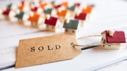 Brown SOLD stamped text label tag with miniature houses on wooden white board. Conceptual image. Selective focus.