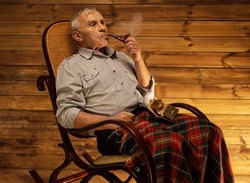 Senior man with smoking pipe sitting on rocking chair in homely wooden interior