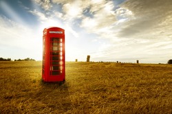 Old-fashioned traditional red telephone booth or public payphone standing in an open deserted field in evening light