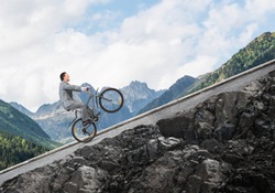 Businessman riding uphill by bike. Nature landscape with copy space. Man in business suit riding bicycle on mountain road. Cyclist popped wheelie on background of blue sky. Healthy lifestyle