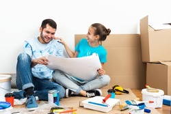 Happy couple sitting on floor with construction blueprint. Home remodeling and house interior redesign. Construction tools and materials lying on floor. Young family studies renovation project of flat