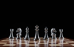 Shiny steel chess figures standing on wooden chessboard. Intellectual duel and tactical battle symbol. Strategy planning and corporate leadership concept. Silver metal chess pieces on black background