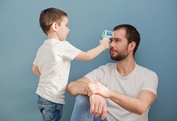 toddler boy is measuring his father's body temperature using infrared thermometer on a blue background
