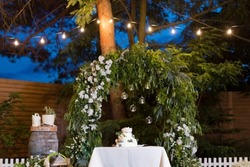 White cake with cream and flowers decoration stand on the table near green wedding arch, light bulb garland and candles