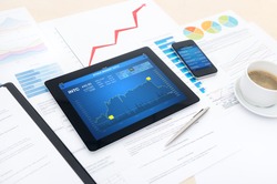Modern business workplace with stock market data application on a digital tablet, mobile banking interface on a smartphone and some papers with charts, graphs and numbers on a desktop.