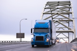 Blue big rig powerful excellent for long-distance industrial transportation semi truck with turn on headlight transporting semi trailer running on arched Fremont bridge in Portland