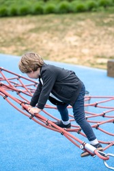 Little boy enthusiastically plays on playground with a stretched rope net to develop the child's balance and motor skills giving him the opportunity to overcome obstacles and achieve victorious goals