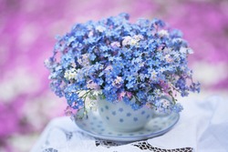 A bouquet of forget-me-not flowers in a cup and saucer on a table with a tablecloth, against a blurred white-pink background of a colorful flower bed.